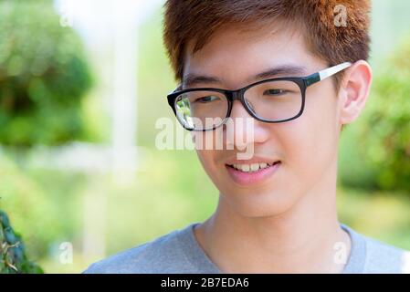 Closeup face Asian handsome teenage boy wearing glasses, Portrait cheerful hipster young man smile with a happy at park on green nature background in Stock Photo