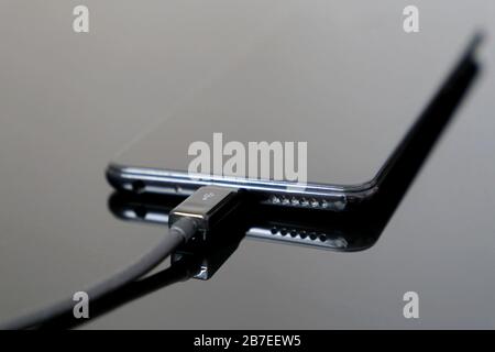 Black smartphone is charging on a dark glass surface. Mobile phone with USB data cable Stock Photo