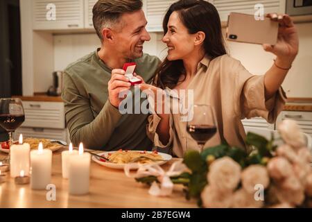 Image of happy caucasian couple taking selfie photo with engagement ring while having romantic candlelight dinner at home Stock Photo