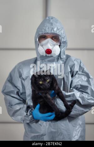 Man in a protective suit, goggles, face mask and disposable gloves stands and holds a black domestic cat on his hands. Fighting the crown with a vir Stock Photo