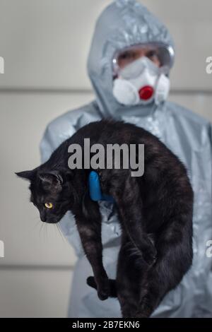 Man in a protective suit, goggles, face mask and disposable gloves stands and holds a black domestic cat on his hands. Fighting the crown with a vir Stock Photo