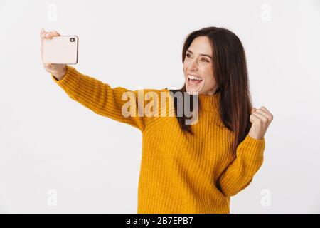 Image of beautiful brunette adult woman wearing sweater smiling and taking selfie photo on cellphone isolated over white background Stock Photo
