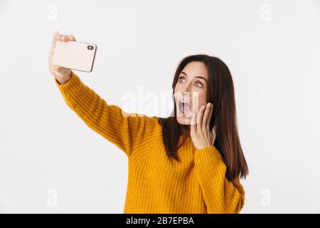 Image of beautiful brunette adult woman wearing sweater smiling and taking selfie photo on cellphone isolated over white background Stock Photo