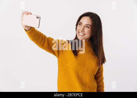 Image of beautiful brunette adult woman wearing sweater smiling and taking selfie photo on cellphone isolated over white background Stock Photo