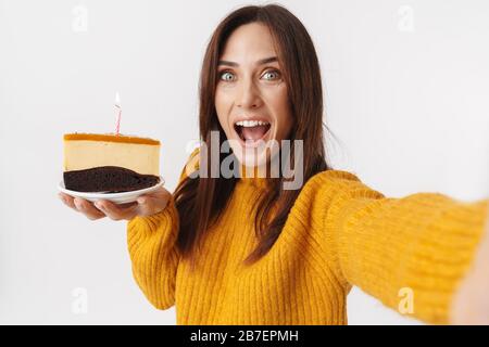 Image of beautiful brunette adult woman wearing sweater holding birthday cake and taking selfie photo isolated over white background Stock Photo