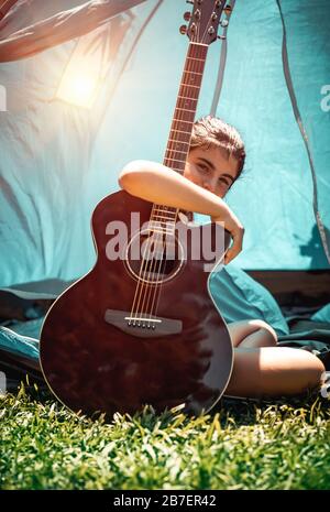 Nice teenager girl sitting on the fresh green grass with guitar, talented child plays a musical instrument, enjoying active time at summer camp Stock Photo