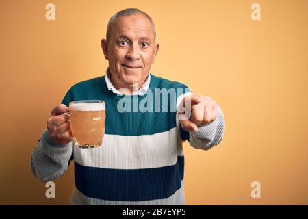 Senior handsome man drinking jar of beer standing over isolated yellow background pointing to you and the camera with fingers, smiling positive and ch Stock Photo