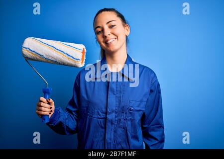 Young beautiful blonde painter woman with blue eyes painting wearing uniform using roller with a happy face standing and smiling with a confident smil Stock Photo