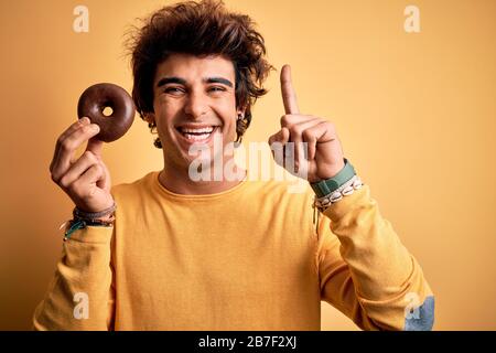Young handsome man holding chocolate donut standing over isolated yellow background surprised with an idea or question pointing finger with happy face Stock Photo
