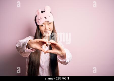 Young asian woman wearing pajama and sleep mask over pink isolated background smiling in love doing heart symbol shape with hands. Romantic concept. Stock Photo