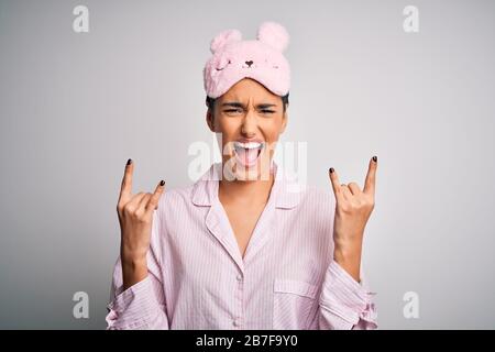 Young beautiful brunette woman wearing pajama and sleep mask over white background shouting with crazy expression doing rock symbol with hands up. Mus