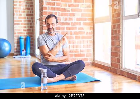 Middle age handsome sportman sitting on mat doing stretching yoga exercise at gym Pointing with hand finger to the side showing advertisement, serious Stock Photo