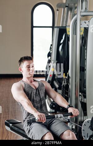 Serious handsome young bodybuilder concentrated on doing exercise in rowing machine in gym Stock Photo