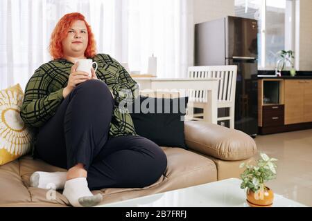 Pensive red haired woman in plaid shirt resting on sofa with big cup of coffee Stock Photo