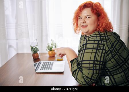 Portrait of smiling red haired overweight woman pointing at screen of opened laptop on her desk Stock Photo