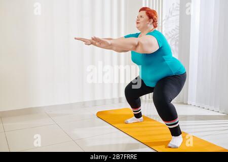 Smiling overweight woman outstretching arms for balance when doing squats at fitness class Stock Photo