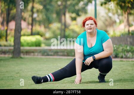 Overweight young woman stretching legs and warming up before training in park Stock Photo