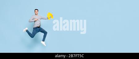 Happy handsome man jumping and pointing to yellow house model isolated on blue banner background with copy space Stock Photo