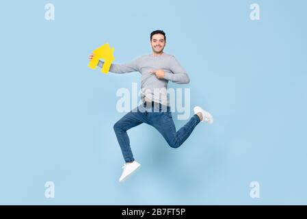 Happy handsome man jumping and pointing to yellow house model isolated on blue studio background Stock Photo