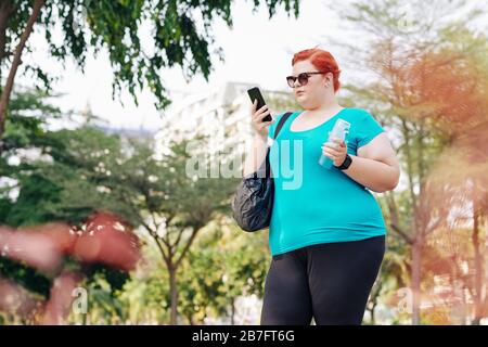 Overweight pretty young woman standing in city park and checking health application on smatphone after training Stock Photo