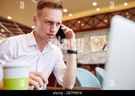 Frowning young businessman reading data on laptop screen and talking on phone with client or coworker Stock Photo
