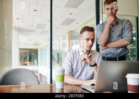 Pensive software developer looking at his colleague coding or making presentation on laptop at office desk Stock Photo