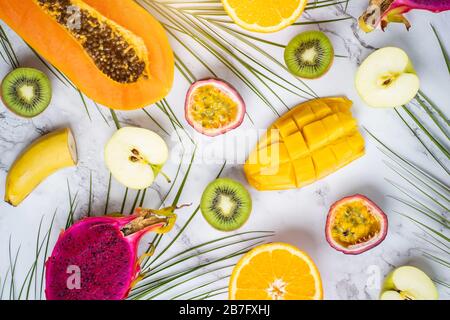 Assortment of different fruits, flat lay, top view. Sliced apple, mango, orange, kiwi, papaya, maracuja, dragon fruit and tropical leaves on white mar Stock Photo