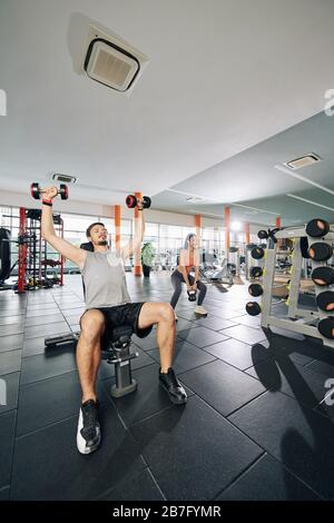 People exercising with heavy weights in gym, young woman doing squats when man lifting dumbbells Stock Photo