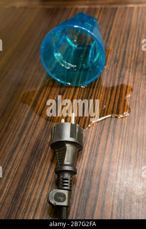 Fallen blue cup with spilled water near an electric cable with a plug on a wooden surface Stock Photo