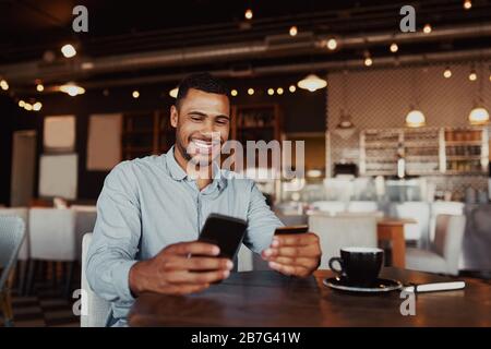 Happy african man holding mobile phone typing card data to make online payment sitting in cafe Stock Photo