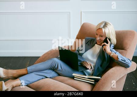 Portrait of smiling mature businesswoman speaking by smartphone while working in lounge chair in office, copy space Stock Photo