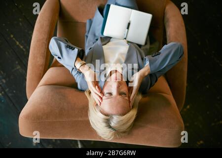 Top view portrait of stressed mature businesswoman rubbing temples suffering from headache while sitting in lounge chair with eyes closed, copy space Stock Photo