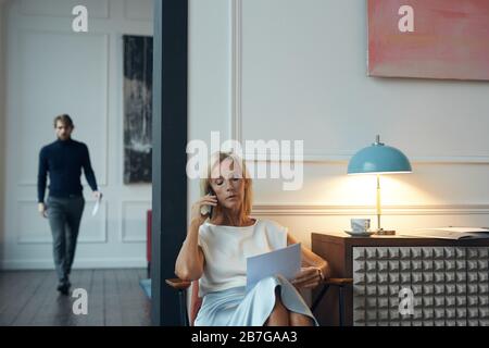 Portrait of successful mature woman speaking by phone and holding documents while sitting in elegant interior with man in background, copy space Stock Photo