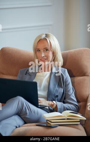 Vertical portrait of successful mature businesswoman smiling at camera while using laptop sitting in lounge chair in office Stock Photo