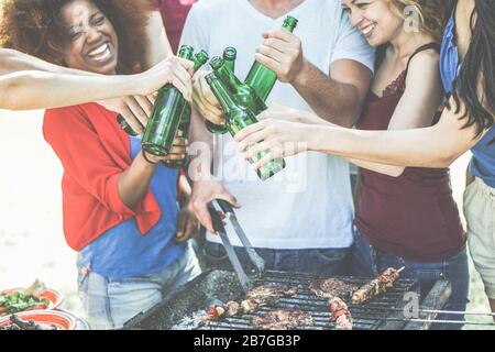 Group of happy diverse culture friends cheering with beer bottles at barbecue party - Young people having fun drinking nd eating together - Friendship Stock Photo