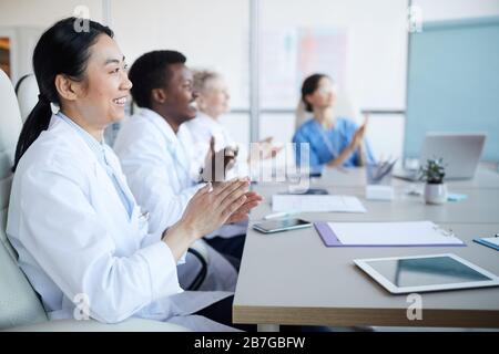 Side view at multi-ethnic group of doctors applauding while sitting at table during medical conference, focus on smiling Asian woman in foreground, co Stock Photo