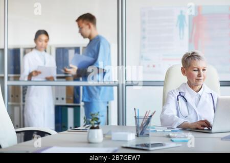 Portrait of mature female doctor using laptop while working in office with other people in background, copy space Stock Photo