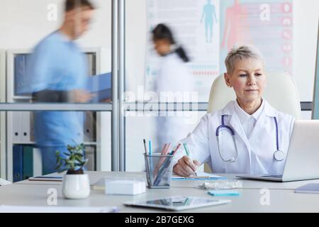 Portrait of senior female doctor using laptop while working in office with other people in background, copy space