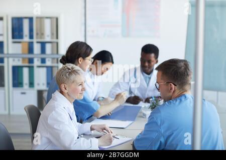 Side view at group of doctors sitting round table during medical council or conference in clinic, copy space Stock Photo