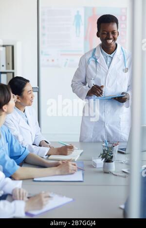 Portrait of young African-American doctor giving speech while standing at table during medical council or conference in clinic, copy space Stock Photo