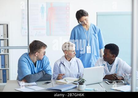 Multi-ethnic group of doctors communicating while sitting at desk with laptop in clinic office interior, copy space Stock Photo