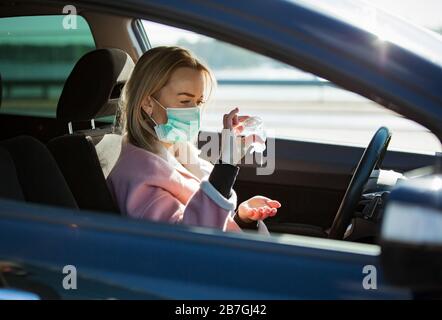 Woman in protective mask sitting in a car on road, using hand sanitizer. Safe traveling. Stock Photo