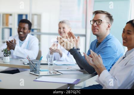 Multi-ethnic group of doctors applauding while sitting at table during medical conference, focus on mature man in foreground, copy space Stock Photo