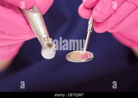 Close-up dentist's hands in a pink latex gloves with mouth mirror and high-speed dental handpiece. Medical tools concept. Shallow depth of fiel. Stock Photo