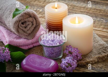 Terry towels, soap for bathroom procedures, burning candles and lilac flowers on sackcloth and old wooden boards. Spa products and accessories. Stock Photo
