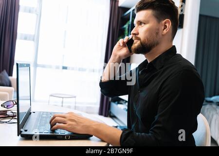 Young bearded man working remotely with a laptop and talking on his cell phone in a modern room. Digital nomad concept Stock Photo