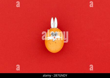 Brown easter egg with funny bunny ears and cartoon face on red background Stock Photo