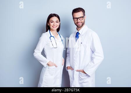 Two best smart professional smiling doctors workers in white coats holding their hands in pockets and together standing against gray background Stock Photo