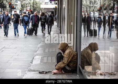 Homeless man sits against a shop window whilst in the distance The Salvation Army Band play music to passers by, Oxford Street, London, UK Stock Photo