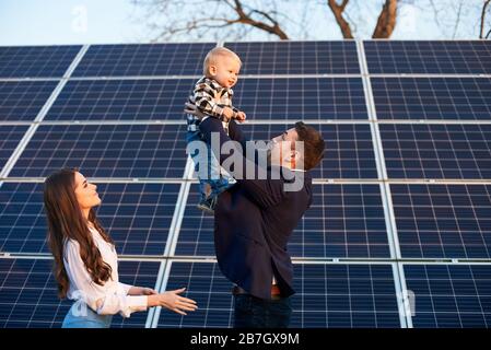 Young man throws up a small child on a background of solar panels under a blue sky. A beautiful girl with long hair is standing nearby. Man in a jacket. Solar energy concept image Stock Photo